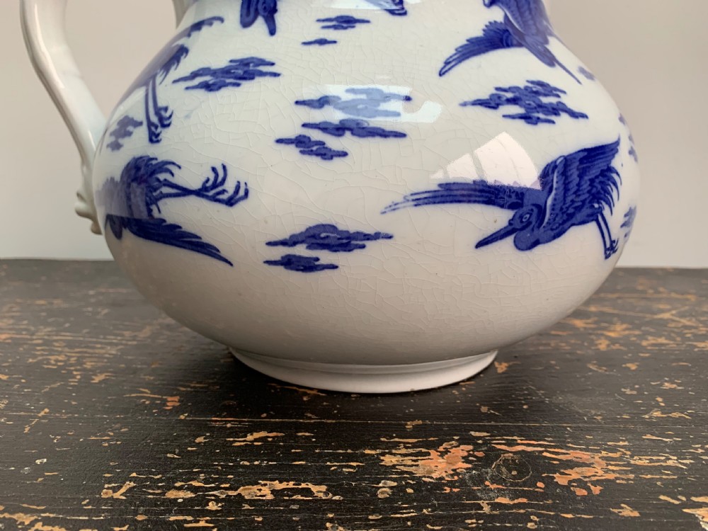 A bulbous white ceramic jug featuring transfer-printed blue flying cranes, a crackle glaze finish, and a moulded handle. View V