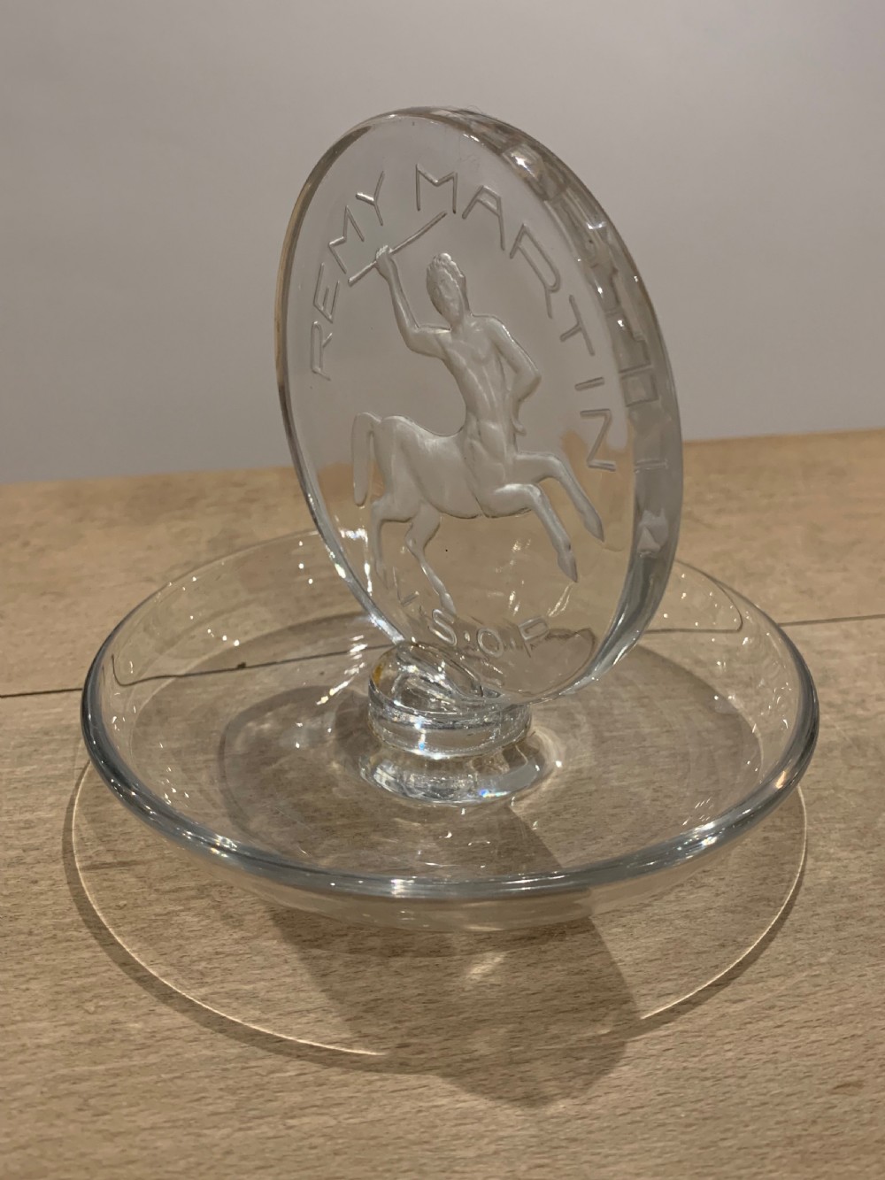 A clear glass Art Deco circular ashtray featuring an upright circular medallion with a frosted relief depicting a centaur holding a javelin above the lettering REMY MARTIN V.S.O.P. View III