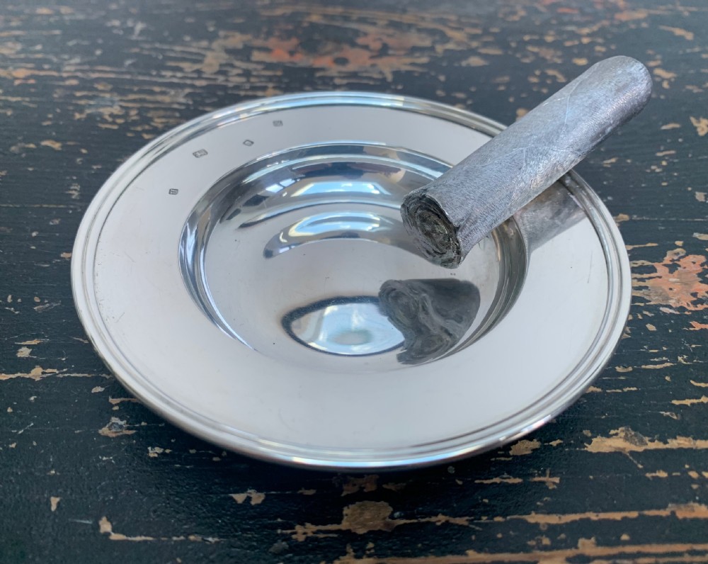 A circular solid silver ashtray featuring a realistic silver trompe loeil cigar resting across its moulded rim.