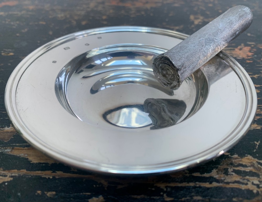 A circular solid silver ashtray featuring a realistic silver trompe loeil cigar resting across its moulded rim. View II