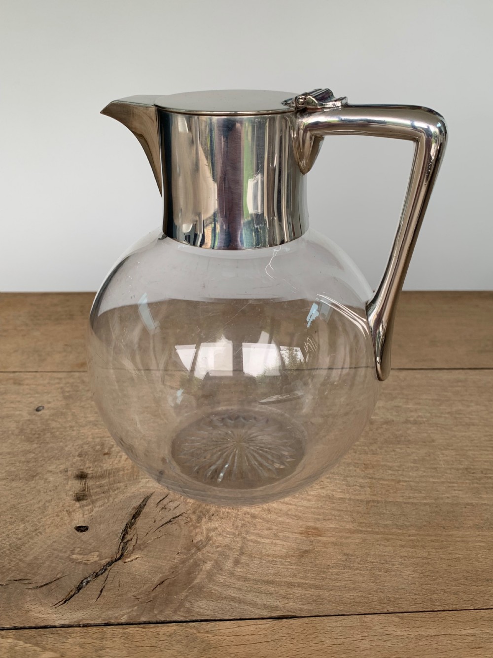 A clear spherical glass claret jug featuring a silver-plated collar, flat hinged cap, angular handle, and a cut starburst pattern on the base.