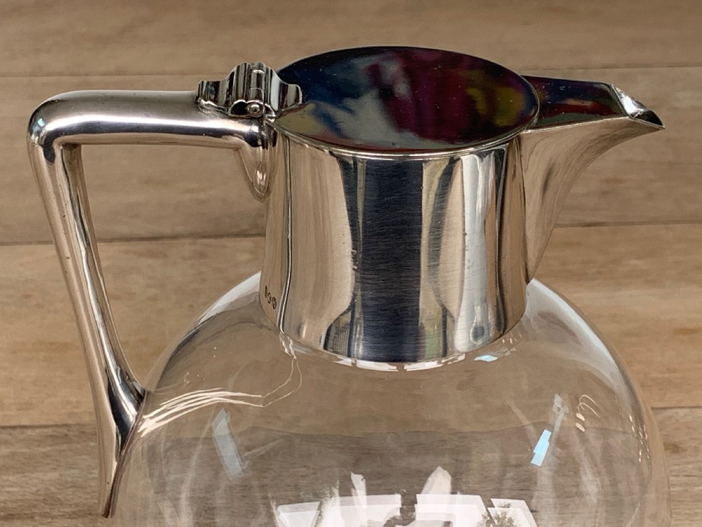 A clear spherical glass claret jug featuring a silver-plated collar, flat hinged cap, angular handle, and a cut starburst pattern on the base. View III