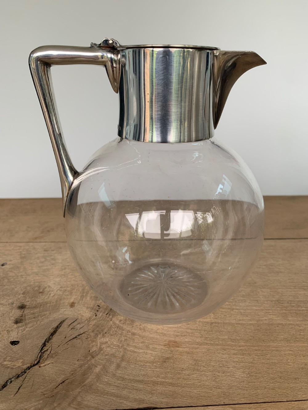 A clear spherical glass claret jug featuring a silver-plated collar, flat hinged cap, angular handle, and a cut starburst pattern on the base. View V