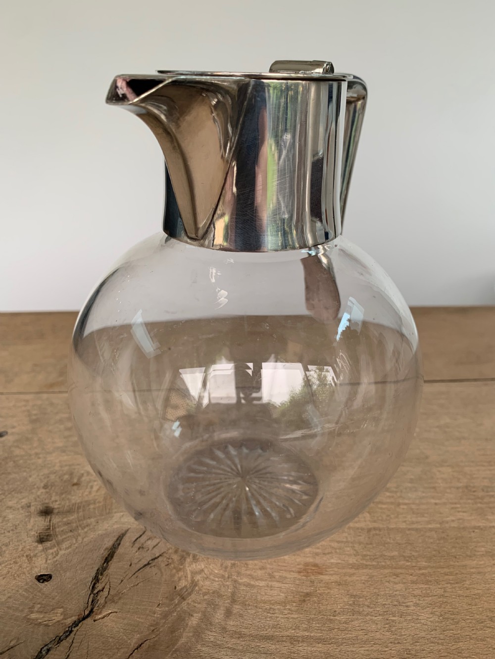 A clear spherical glass claret jug featuring a silver-plated collar, flat hinged cap, angular handle, and a cut starburst pattern on the base. View VI