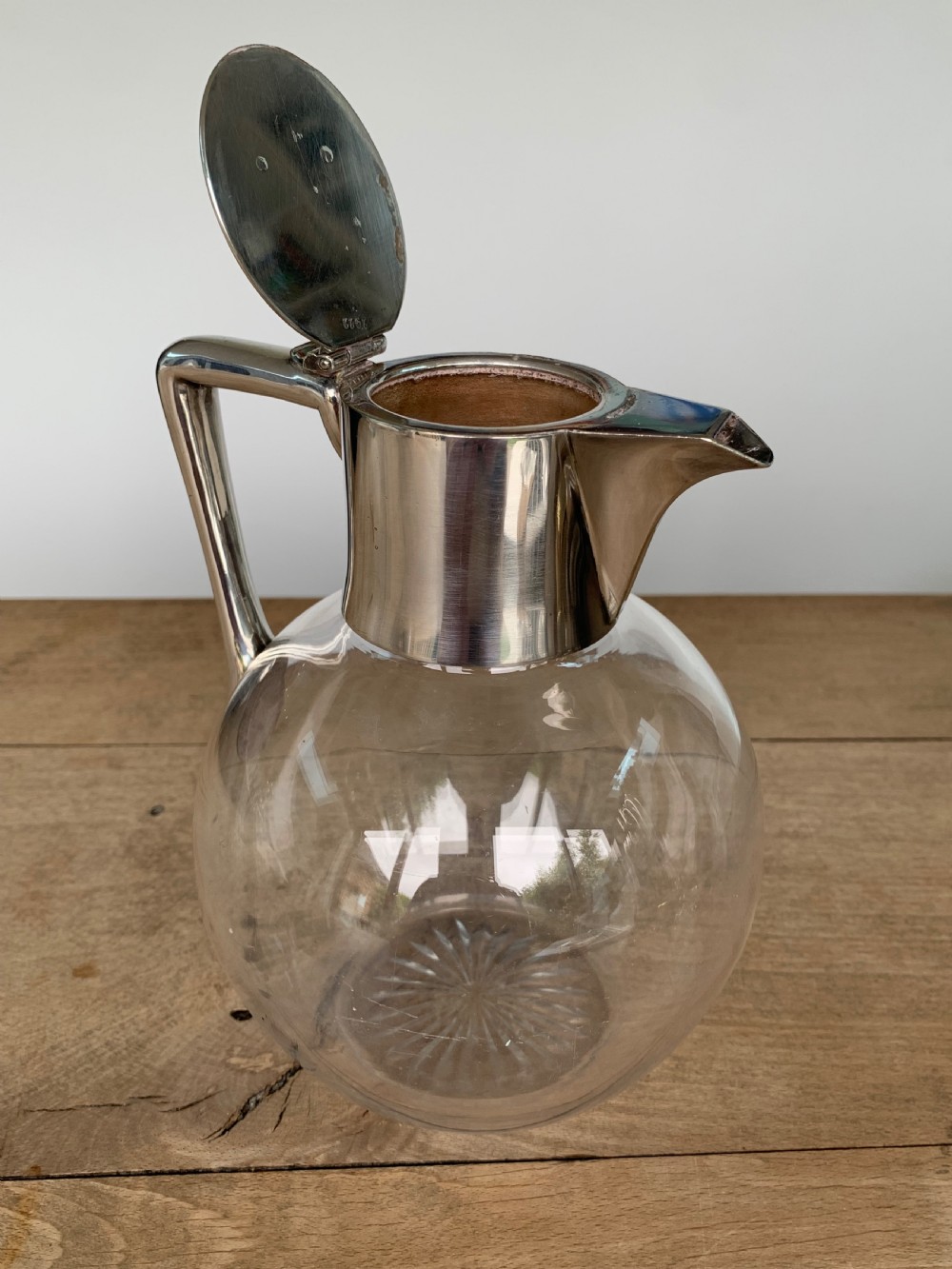 A clear spherical glass claret jug featuring a silver-plated collar, flat hinged cap, angular handle, and a cut starburst pattern on the base. View X