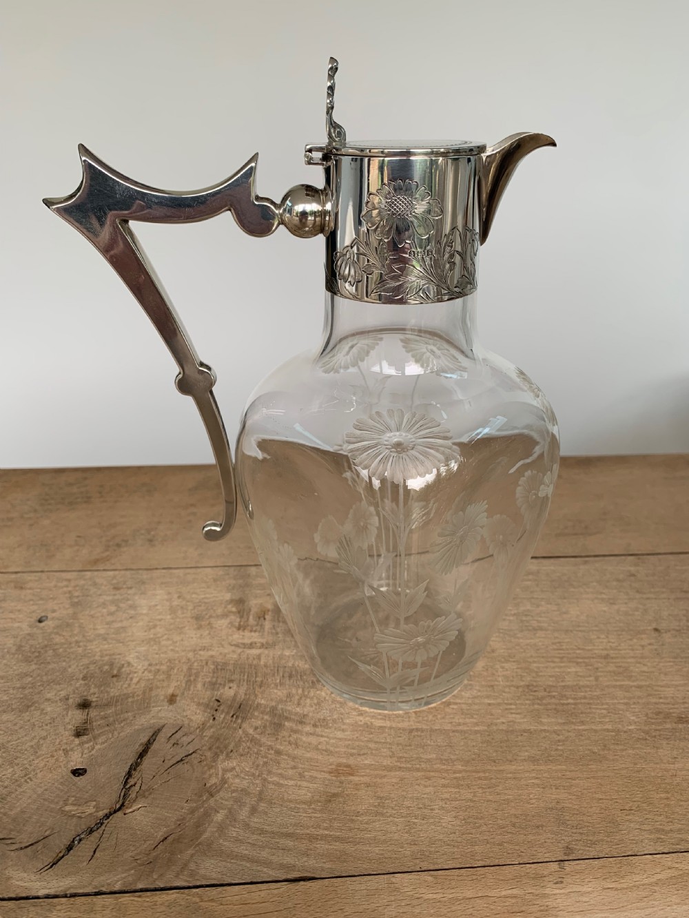 An Edwardian clear glass claret jug featuring wheel-engraved daisy motifs and a hallmarked sterling silver collar with handle.