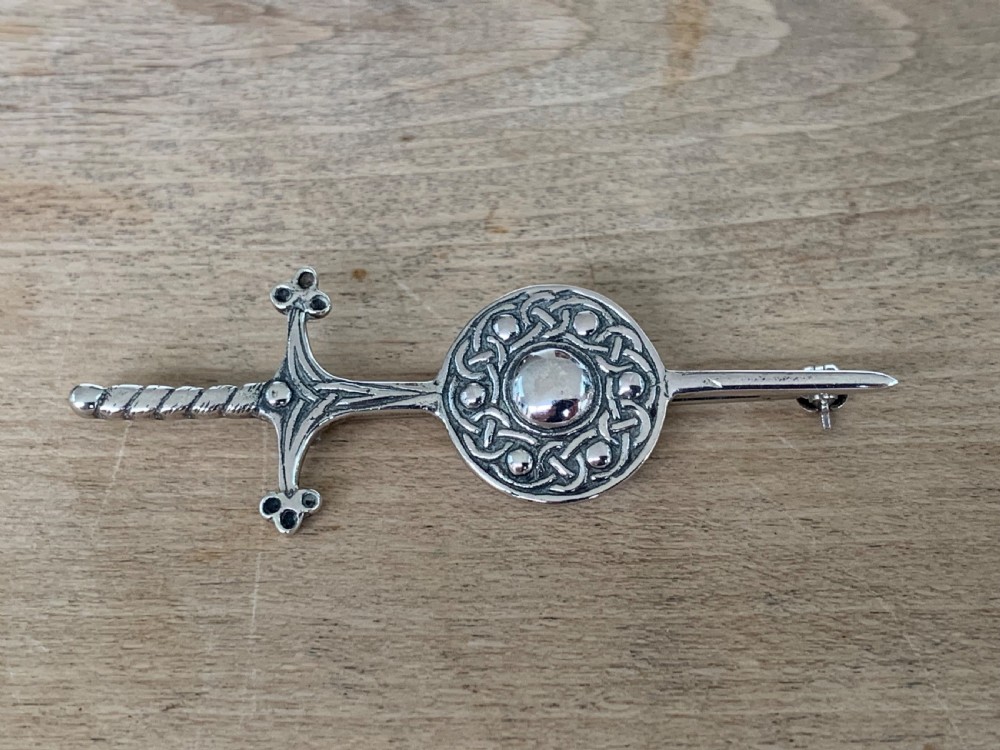 A sterling silver brooch designed in the shape of a sword passing through a circular shield embellished with intricate Celtic knotwork.