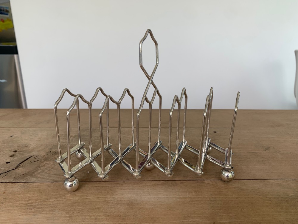 An expandable silver plated Victorian toast rack sits on a wooden surface, showing its collapsible metal frame and central loop handle. View II