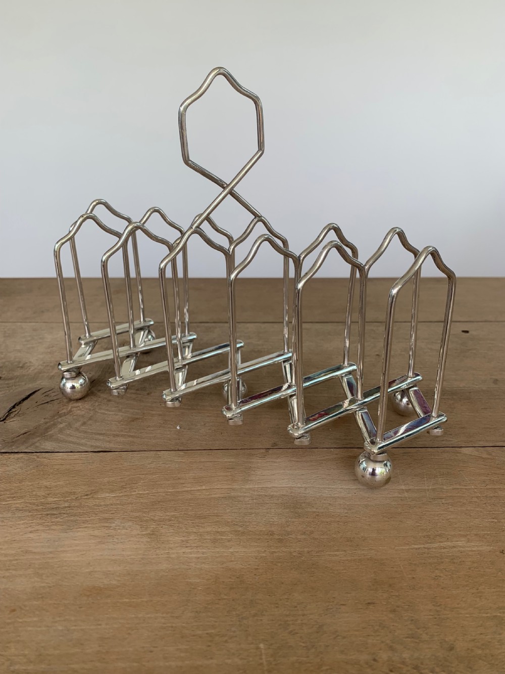 An expandable silver plated Victorian toast rack sits on a wooden surface, showing its collapsible metal frame and central loop handle. View III