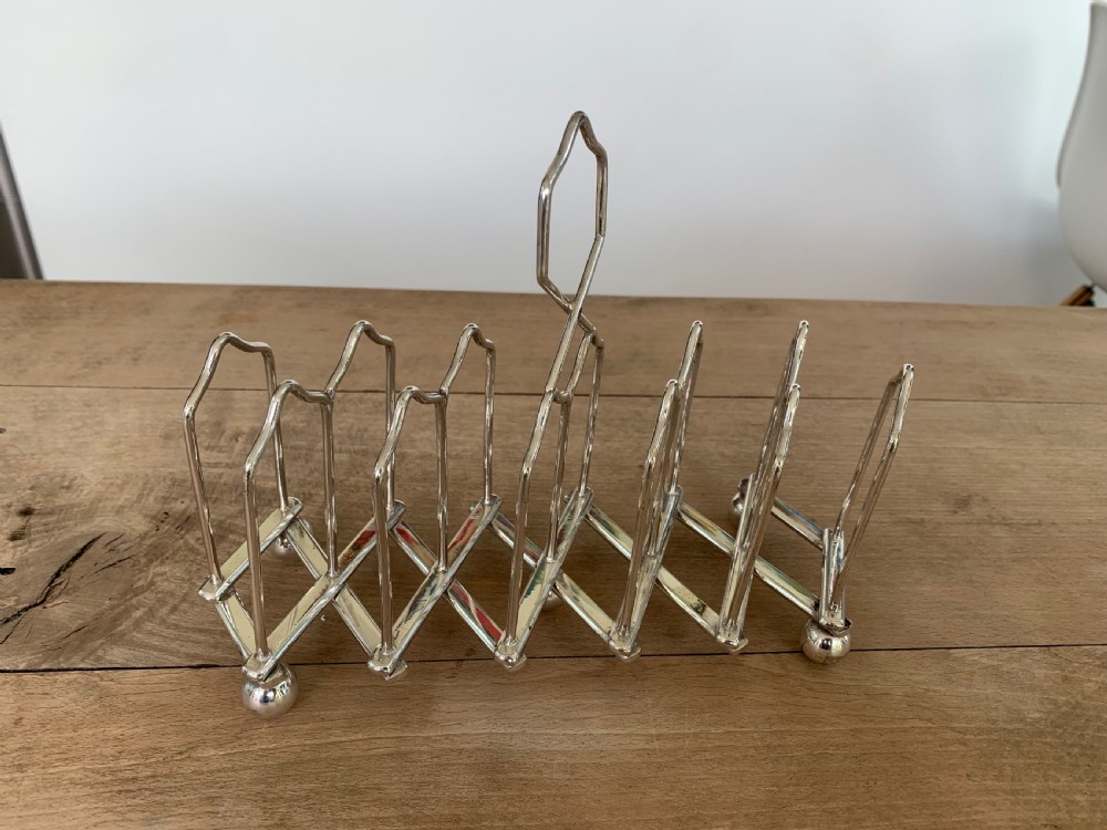 An expandable silver plated Victorian toast rack sits on a wooden surface, showing its collapsible metal frame and central loop handle. View IV