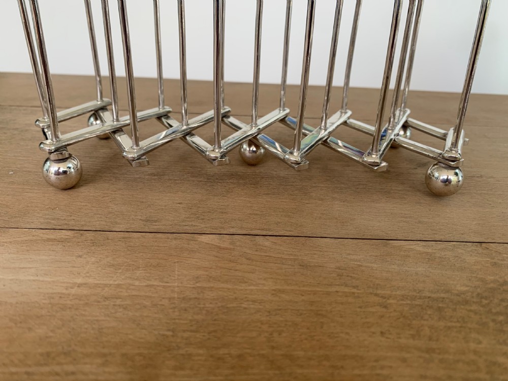 An expandable silver plated Victorian toast rack sits on a wooden surface, showing its collapsible metal frame and central loop handle. View V
