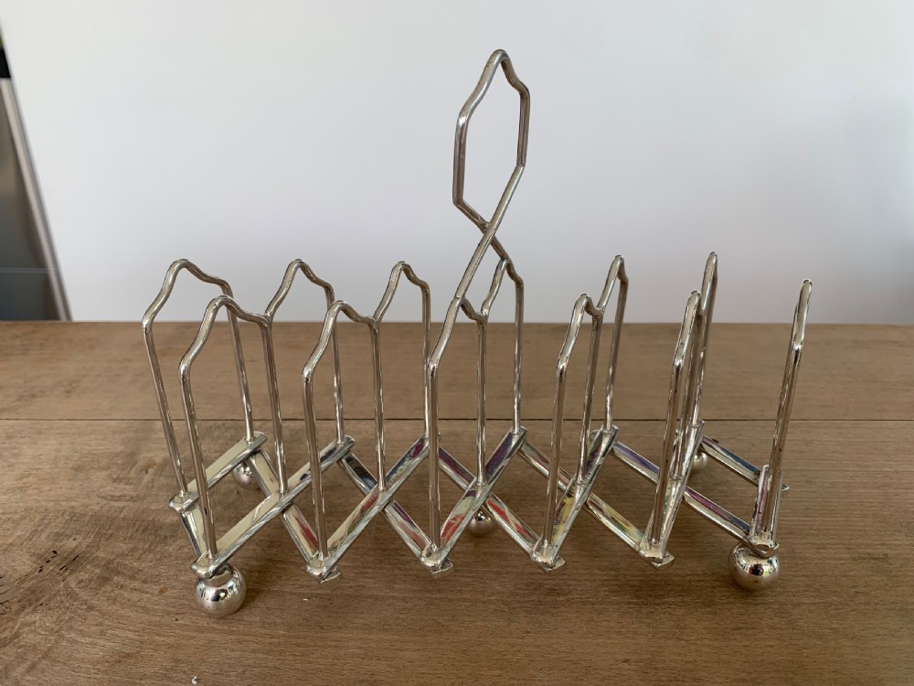 An expandable silver plated Victorian toast rack sits on a wooden surface, showing its collapsible metal frame and central loop handle. View VI
