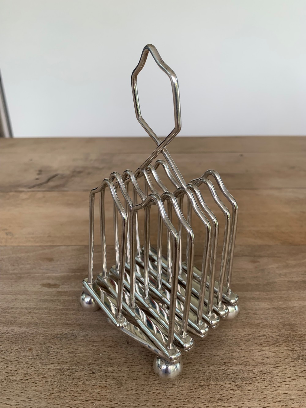 An expandable silver plated Victorian toast rack sits on a wooden surface, showing its collapsible metal frame and central loop handle. View VII