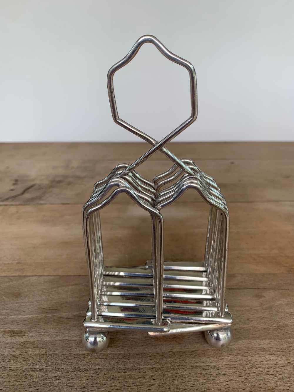 An expandable silver plated Victorian toast rack sits on a wooden surface, showing its collapsible metal frame and central loop handle. View VIII