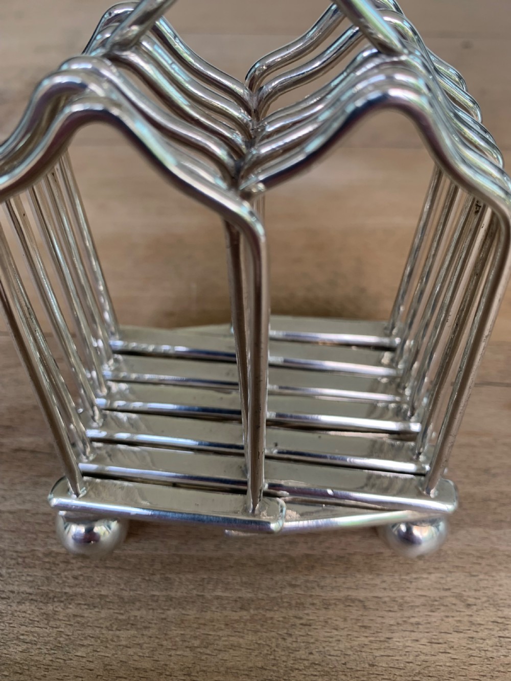 An expandable silver plated Victorian toast rack sits on a wooden surface, showing its collapsible metal frame and central loop handle. View IX