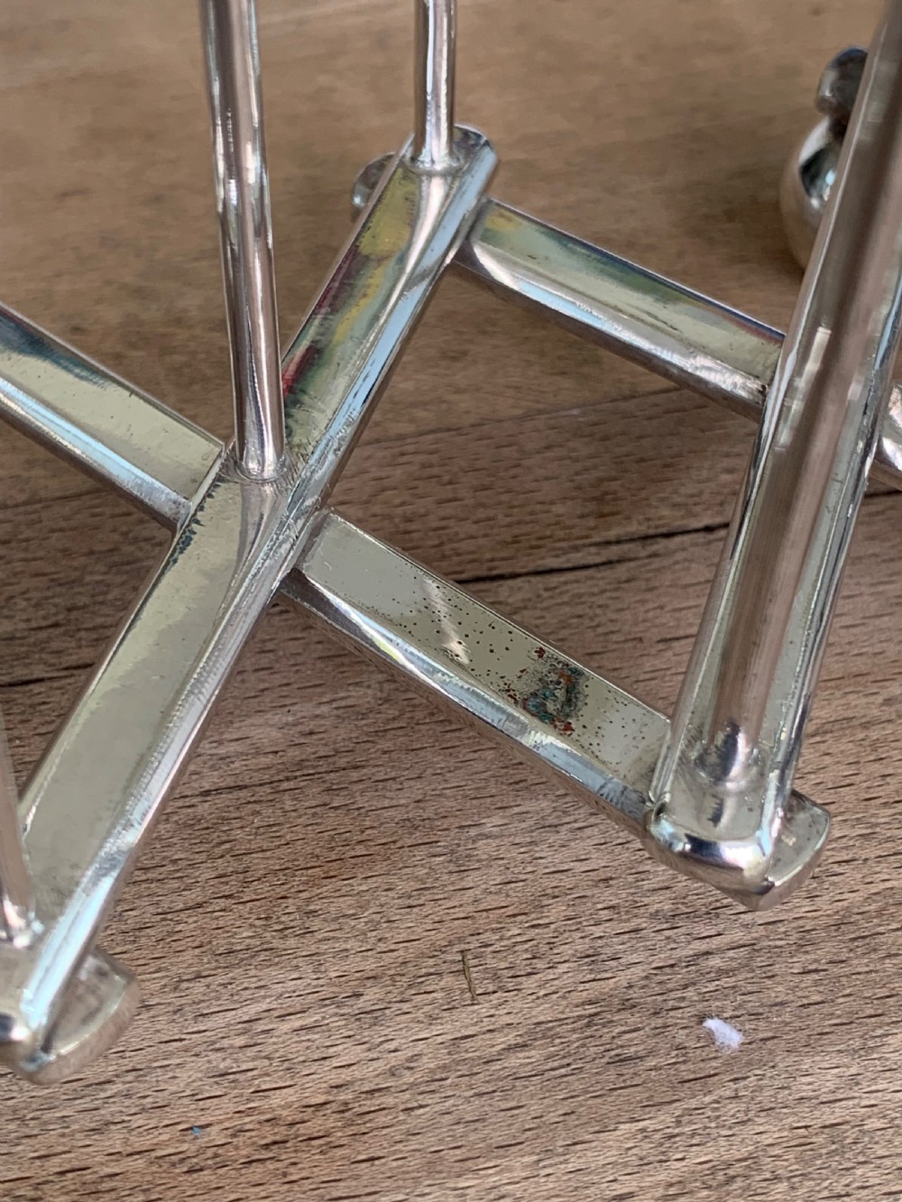 An expandable silver plated Victorian toast rack sits on a wooden surface, showing its collapsible metal frame and central loop handle. View X