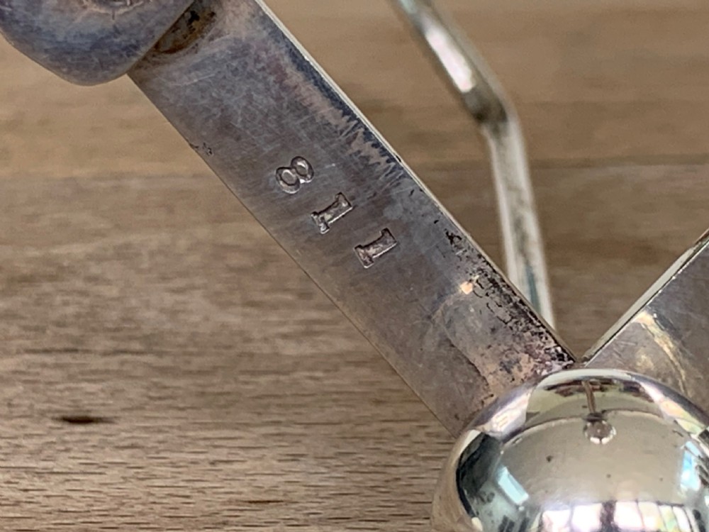 An expandable silver plated Victorian toast rack sits on a wooden surface, showing its collapsible metal frame and central loop handle. View XIII