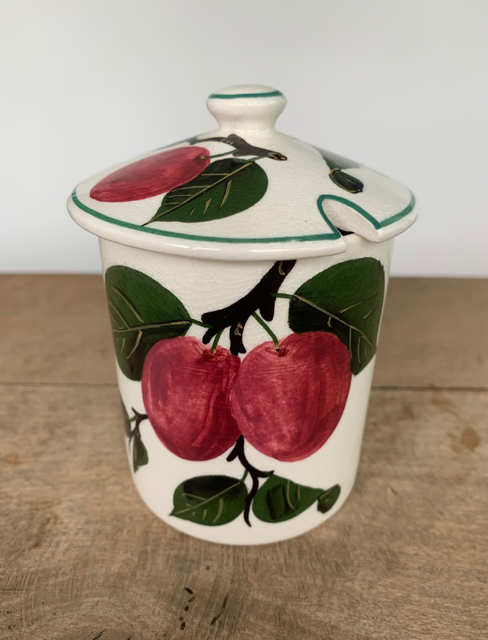 A lidded white ceramic preserve pot decorated with hand-painted red cherries and green leaves, featuring a notch for a spoon.