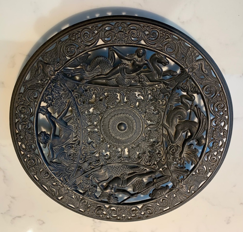 A circular dark charcoal cast iron charger featuring intricate pierced scrollwork and raised classical figures around a central medallion.