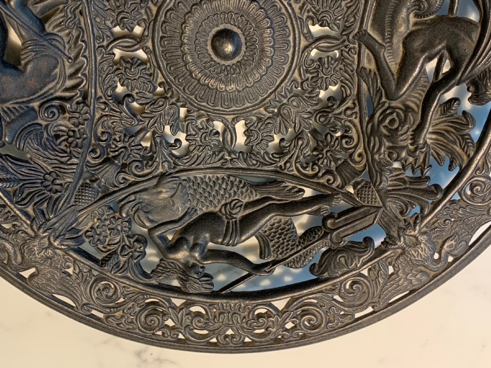 A circular dark charcoal cast iron charger featuring intricate pierced scrollwork and raised classical figures around a central medallion. View II