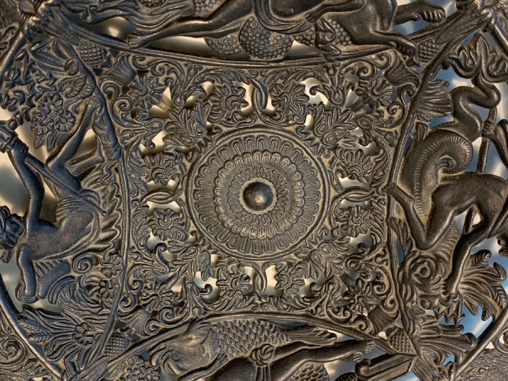 A circular dark charcoal cast iron charger featuring intricate pierced scrollwork and raised classical figures around a central medallion. View III