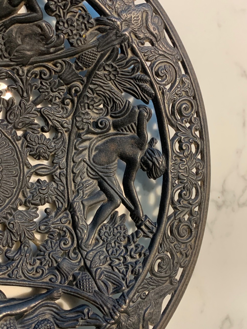 A circular dark charcoal cast iron charger featuring intricate pierced scrollwork and raised classical figures around a central medallion. View IV