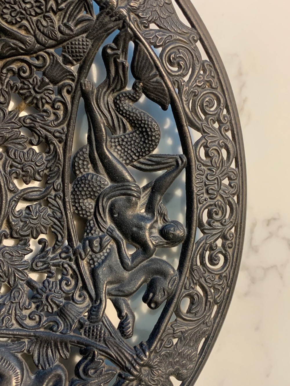 A circular dark charcoal cast iron charger featuring intricate pierced scrollwork and raised classical figures around a central medallion. View V