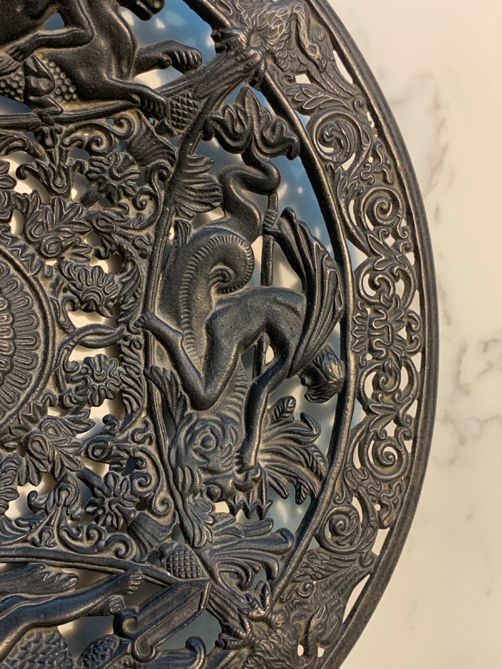 A circular dark charcoal cast iron charger featuring intricate pierced scrollwork and raised classical figures around a central medallion. View VI