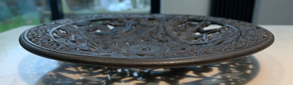 A circular dark charcoal cast iron charger featuring intricate pierced scrollwork and raised classical figures around a central medallion. View VII