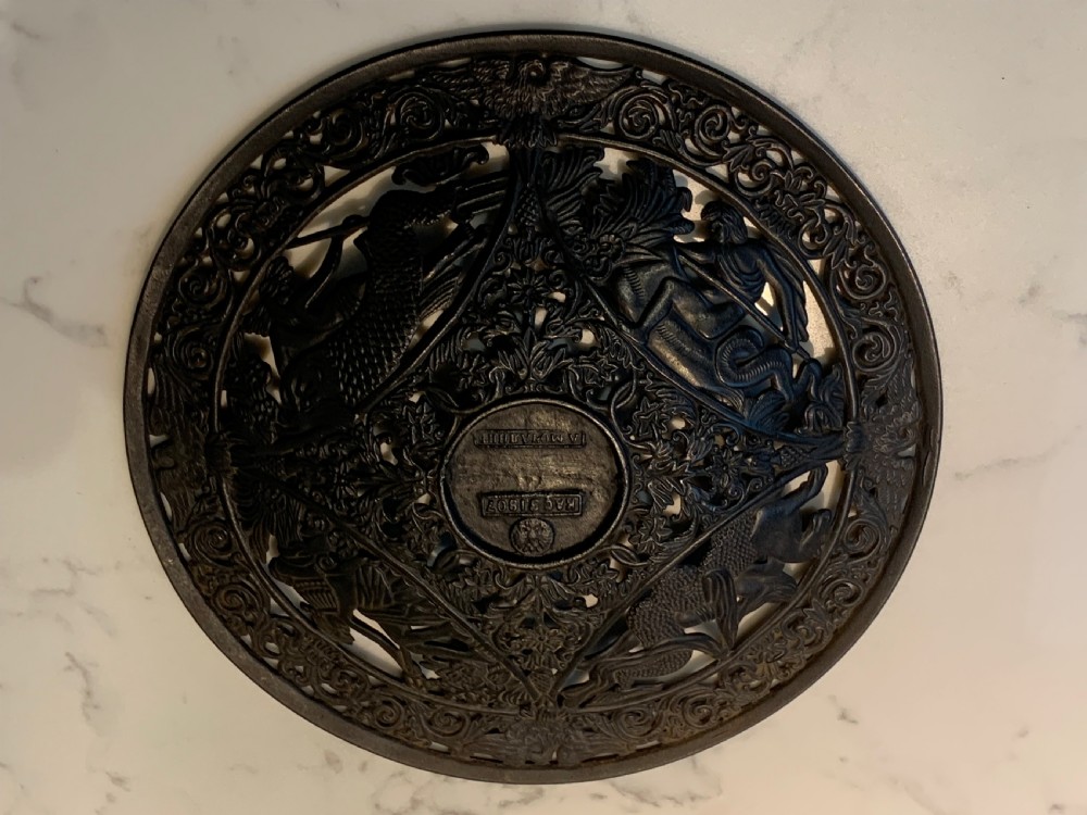 A circular dark charcoal cast iron charger featuring intricate pierced scrollwork and raised classical figures around a central medallion. View VIII