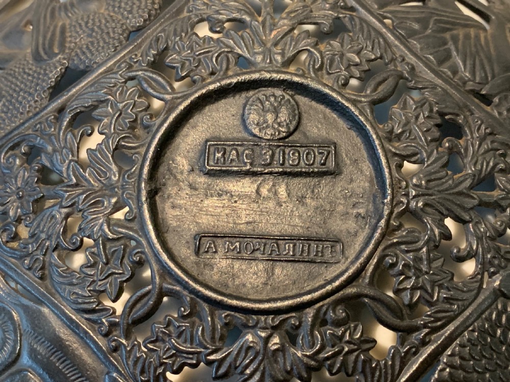 A circular dark charcoal cast iron charger featuring intricate pierced scrollwork and raised classical figures around a central medallion. View IX