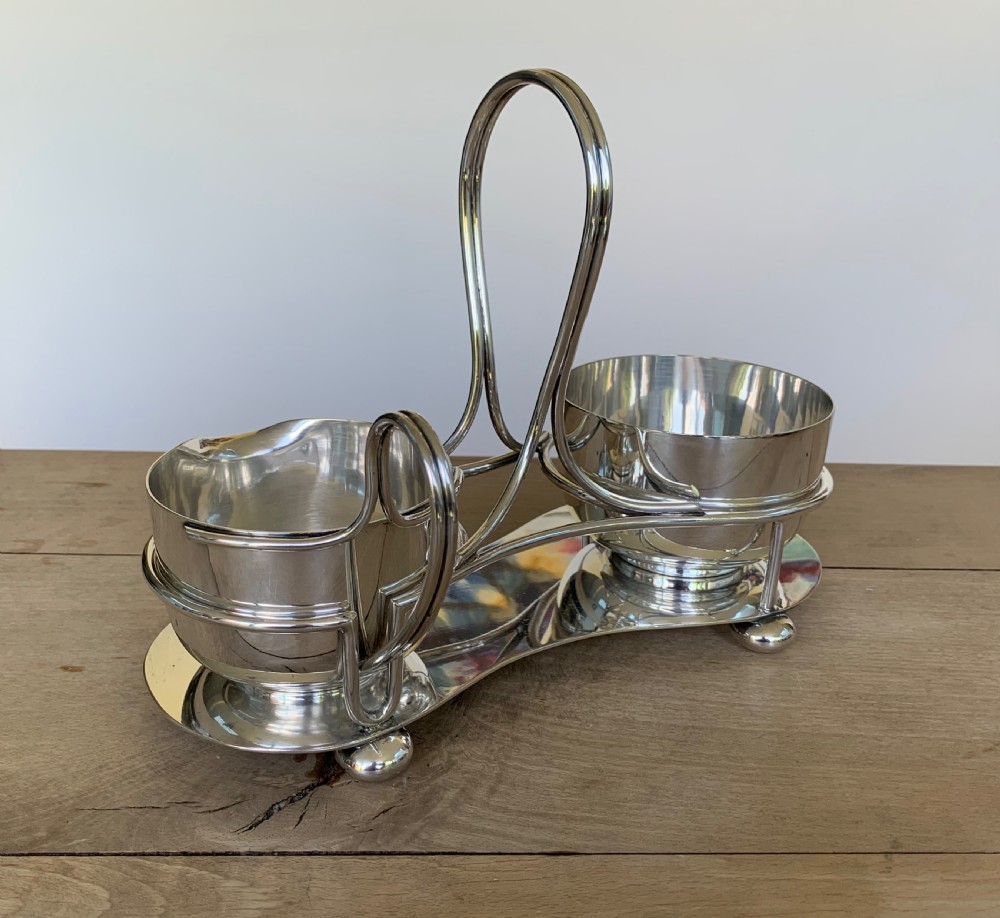 An Art Deco silver-plated sugar bowl and cream jug set within a fitted metal frame featuring a tall loop handle and ball feet.