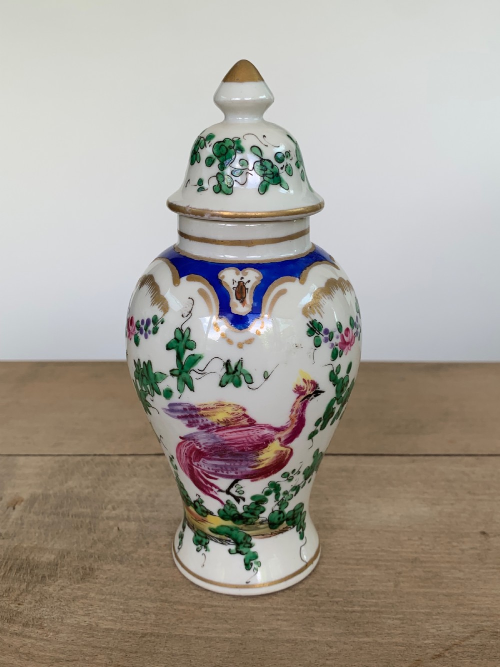 A white lidded porcelain vase decorated with a hand-painted pink exotic bird, green foliage, cobalt blue panels, and gilt trim.