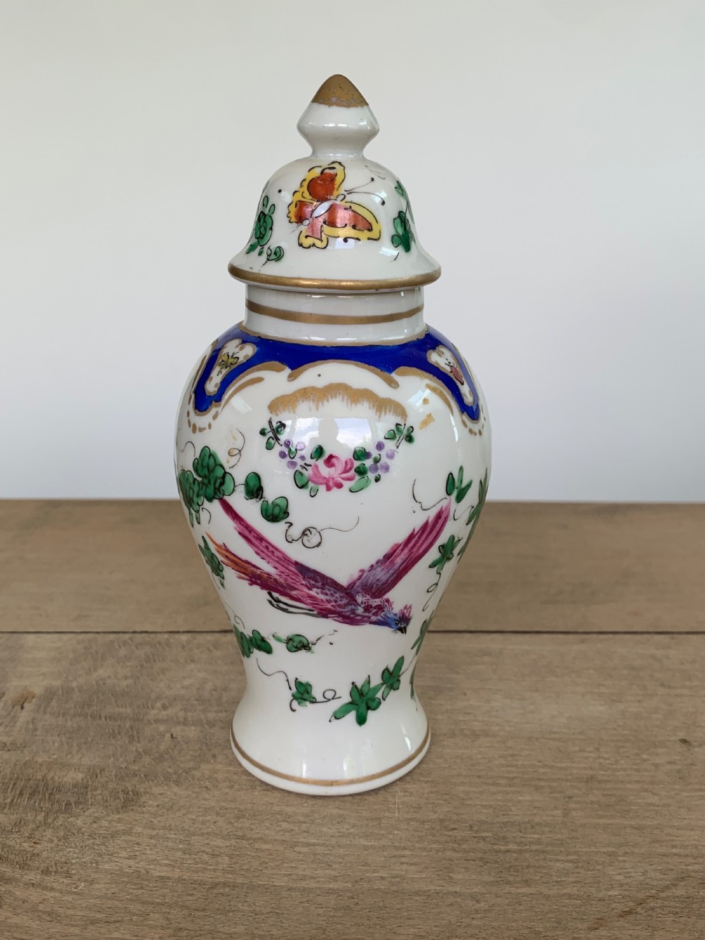 A white lidded porcelain vase decorated with a hand-painted pink exotic bird, green foliage, cobalt blue panels, and gilt trim. View II
