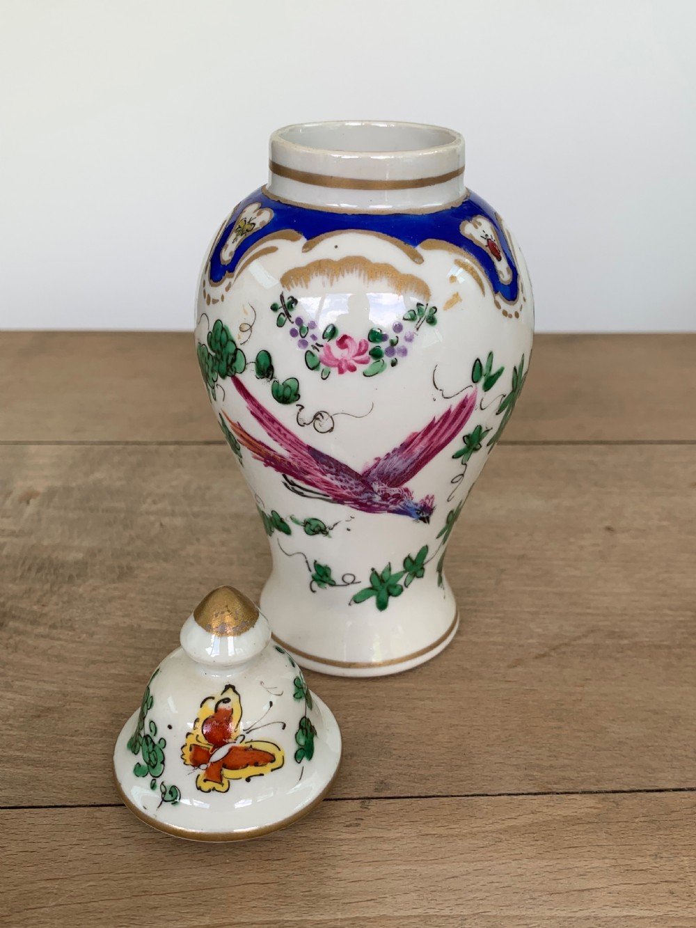 A white lidded porcelain vase decorated with a hand-painted pink exotic bird, green foliage, cobalt blue panels, and gilt trim. View V