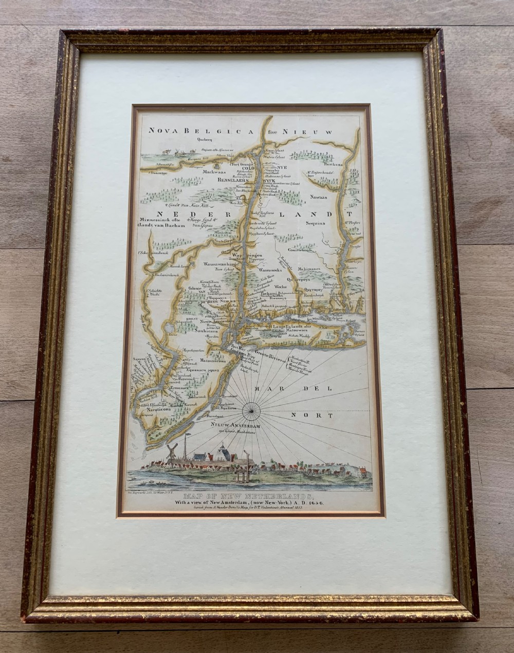 A framed antique map of New Netherlands featuring a detailed compass rose and an inset panorama illustration of early New Amsterdam along the bottom. View II