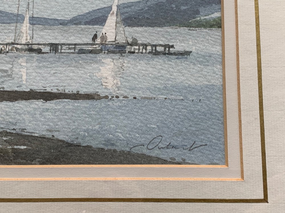 A framed watercolour painting by Stanley Orchart depicting sailboats, jetties, and stone buildings on the shore of Loch Earn under a cloud-filled blue sky. View IV