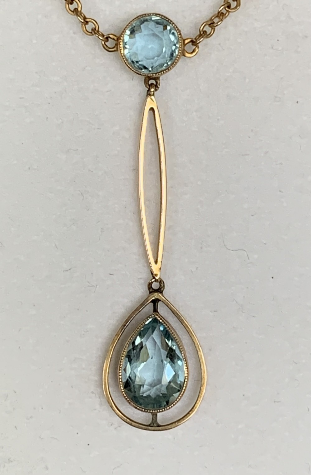 An Edwardian 9ct yellow gold drop necklace featuring a round light blue gemstone linked to a pear-cut gemstone in an openwork frame. View V
