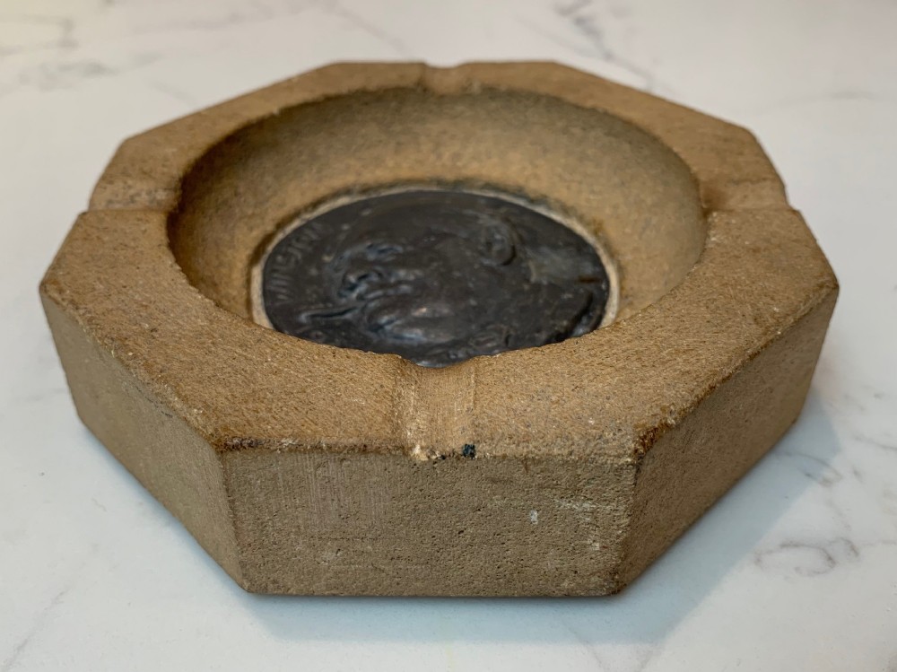 An octagonal buff stone ashtray featuring a central dark metal relief medallion depicting Winston Churchill smoking a cigar.
