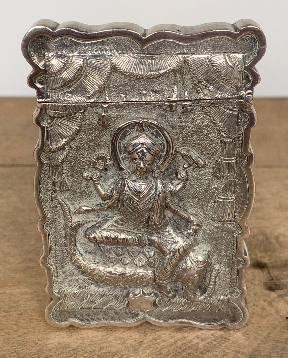 A rectangular silver card case featuring repousse metalwork depicting a multi-armed Hindu deity framed by a scalloped border.