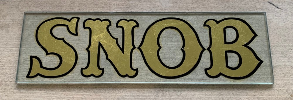A rectangular clear glass plaque displaying the word SNOB in gold leaf lettering outlined in black paint.