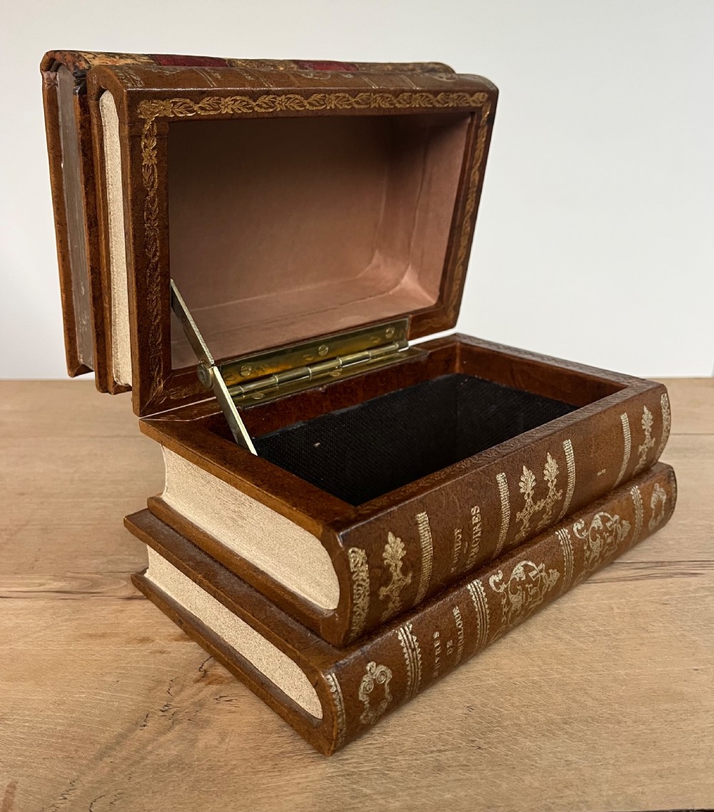 An open vintage warm brown leather box disguised as a stack of two gold-tooled books, featuring a fabric-lined interior with a brass support mechanism.