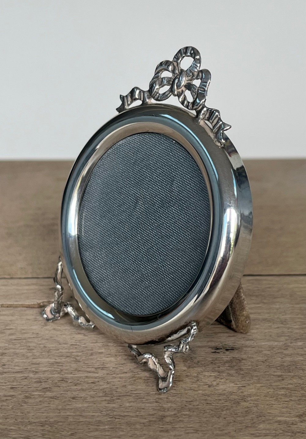 A round sterling silver picture frame featuring an ornate ribbon bow finial at the top and two scrolled support feet at the base.