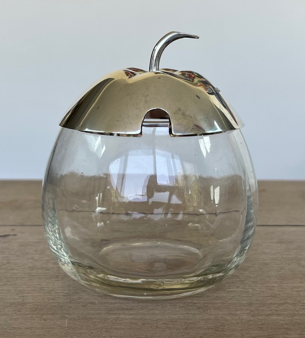 A clear glass preserve pot shaped like an apple with a fitted sterling silver lid featuring a curved stem handle and a spoon notch.