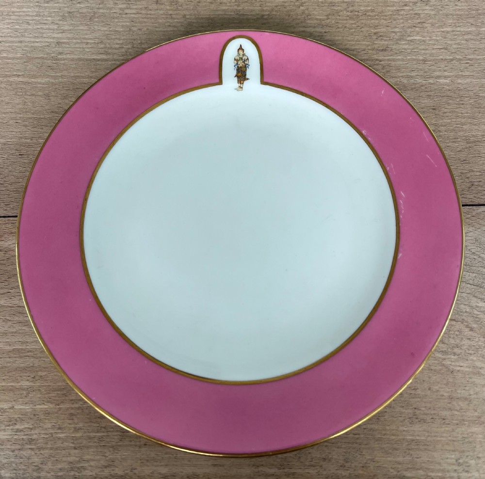 An early 20th-century circular porcelain side plate featuring a broad pink border, gilded rim, white centre, and a small painted Thai figure in a top reserve.