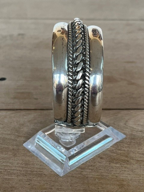 A wide, open-cuff solid silver bracelet featuring a central braided rope design flanked by smooth outer edges and stamped hallmarks. View VI