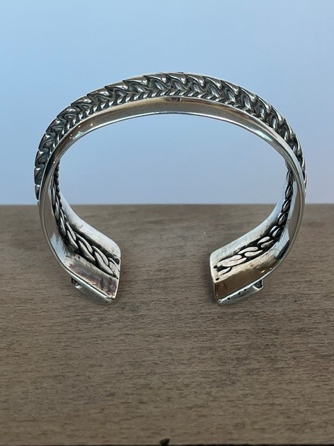A wide, open-cuff solid silver bracelet featuring a central braided rope design flanked by smooth outer edges and stamped hallmarks. View VII