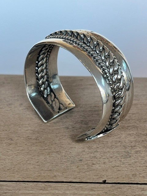 A wide, open-cuff solid silver bracelet featuring a central braided rope design flanked by smooth outer edges and stamped hallmarks. View IX
