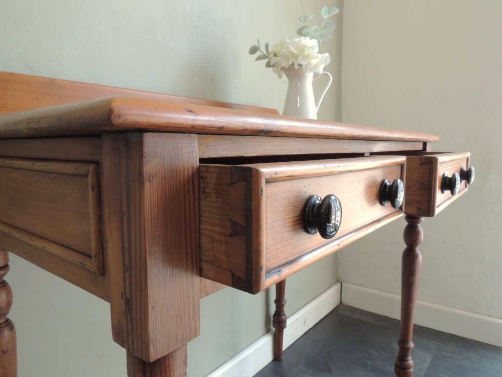 A Victorian solid pine hall table featuring a low gallery backboard, two drawers with dark round knobs, and four turned legs. View V