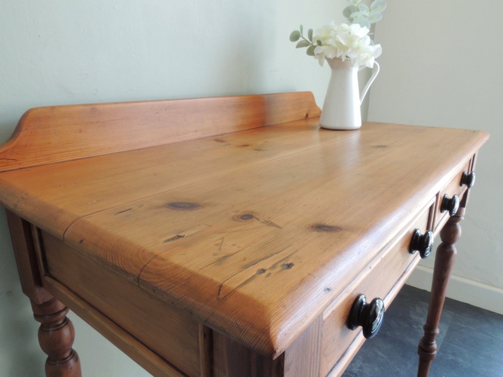 A Victorian solid pine hall table featuring a low gallery backboard, two drawers with dark round knobs, and four turned legs. View VI